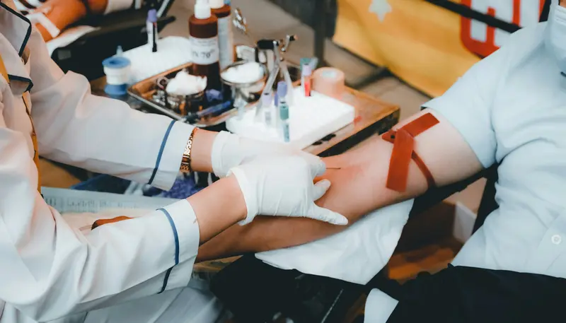 Un profesional sanitario, provisto de guantes, se dispone a extraer sangre del brazo de una persona a la que se le ha colocado un torniquete, con los tubos de muestra y el material necesario en una bandeja situada cerca.