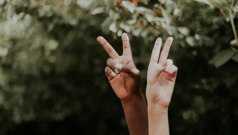 Dos manos levantadas, de distintos tonos de piel, forman el signo de la paz sobre un fondo verde difuminado y frondoso.