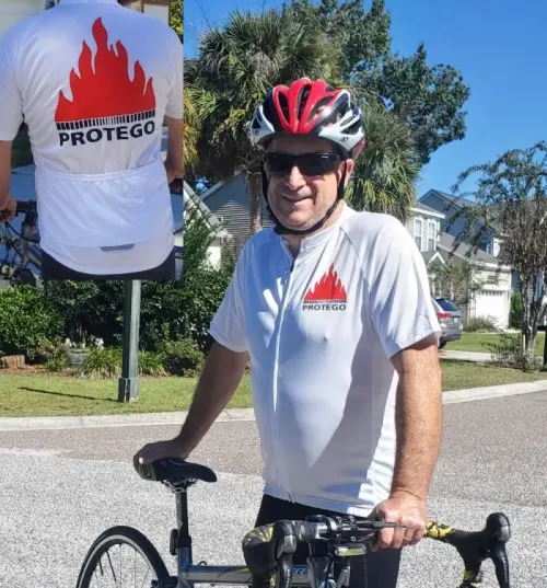 Un ciclista con casco rojo posa junto a su bicicleta de carretera, vestido con un maillot blanco de PROTEGO, en cuya espalda se aprecia un recuadro con el gran logotipo de PROTEGO en forma de llama.