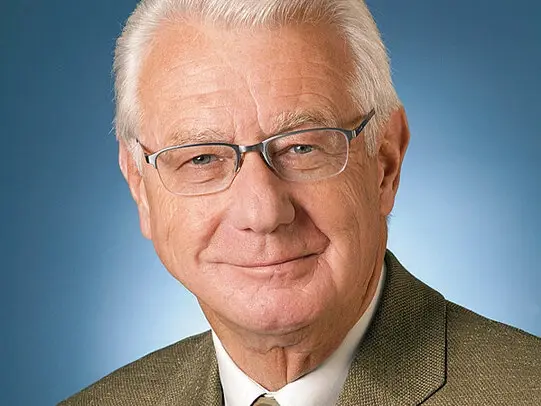 A professional headshot of an elderly man with white hair and glasses, wearing a light shirt, tie, and brown blazer, smiling slightly against a blue studio background.