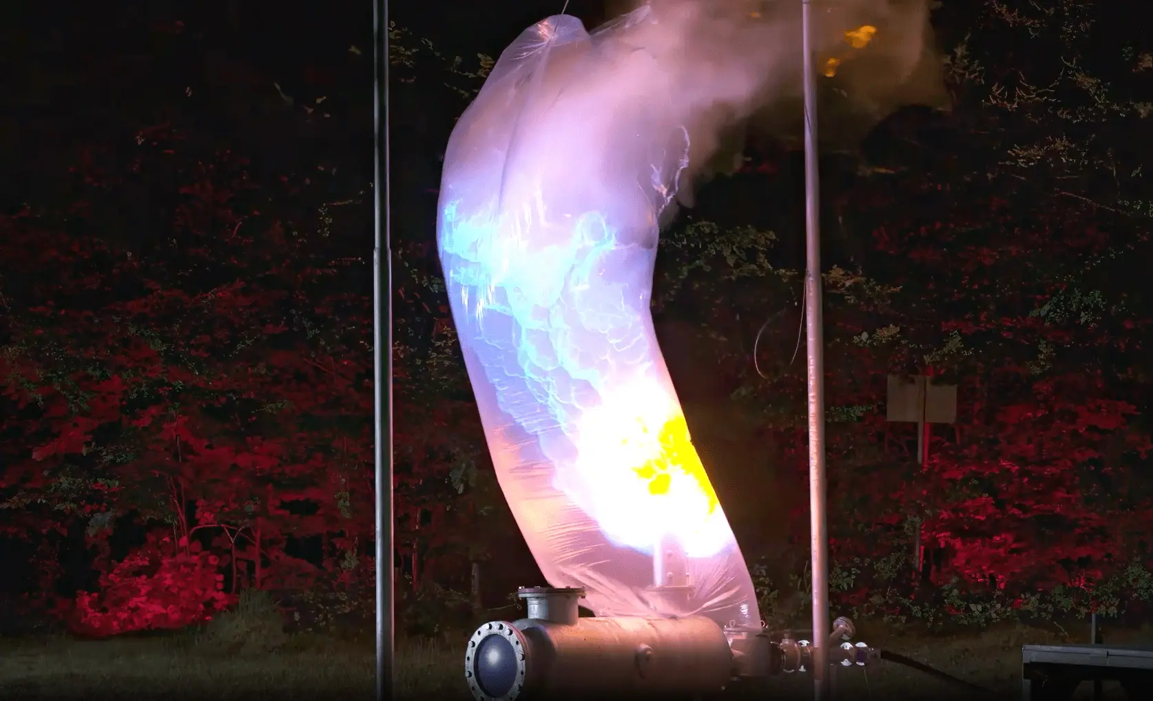 Nighttime industrial test showing a curved pipe releasing a large, brightly glowing flame or ignition plume inside a translucent protective sleeve, illuminated against a wooded background.