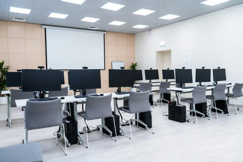 Uma sala de informática vazia, com fileiras de mesas, monitores de computador e uma grande tela de projeção na frente.