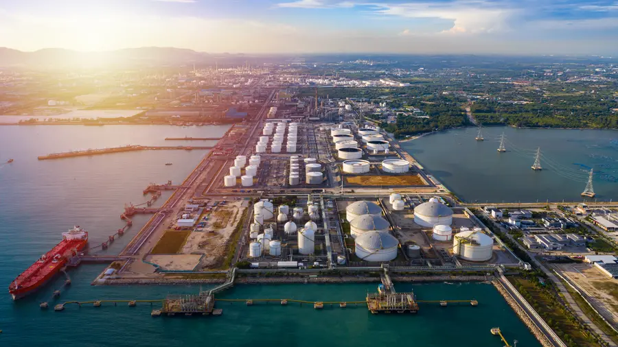 Vista aérea de una refinería industrial costera con hileras de tanques de almacenamiento blancos, muelles con un buque cisterna rojo y canales que se extienden hasta el horizonte al atardecer.
