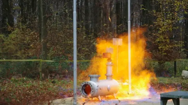 Outdoor industrial test showing a pipeline-mounted safety device surrounded by a large yellow-orange flame plume, set against a wooded background.