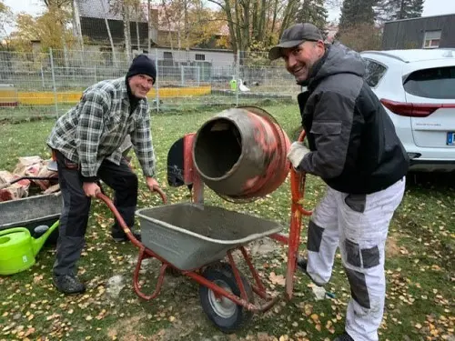 Zwei Arbeiter lächeln im Freien neben einer Schubkarre und einem Betonmischer, umgeben von Laub und einem geparkten Auto.