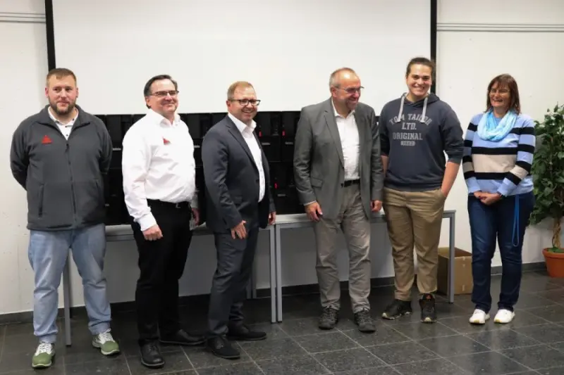 Seis pessoas posam sorridentes para uma foto em grupo numa sala de aula, em frente a uma mesa repleta de malas pretas com equipamentos e uma tela de projeção.