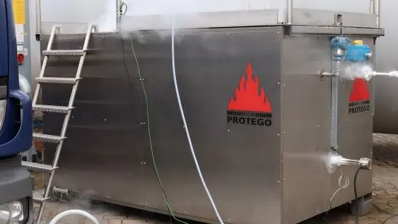Stainless steel industrial test container with Protego logos, external piping, and a ladder, set up outdoors with hoses connected and light vapor visible.