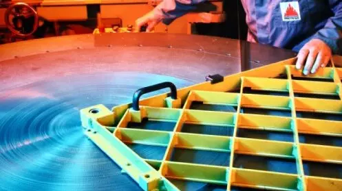 A worker measures a large circular metal plate beside a yellow grid-like frame in an industrial workshop.