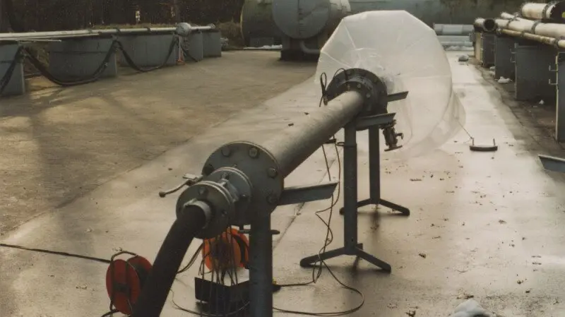 An outdoor test setup shows a long metal pipe on stands with cables and a clear plastic bag attached at the far end.