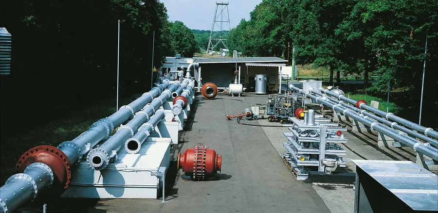 An outdoor industrial test site with multiple large pipelines, valves, and equipment arranged along a paved yard bordered by trees.