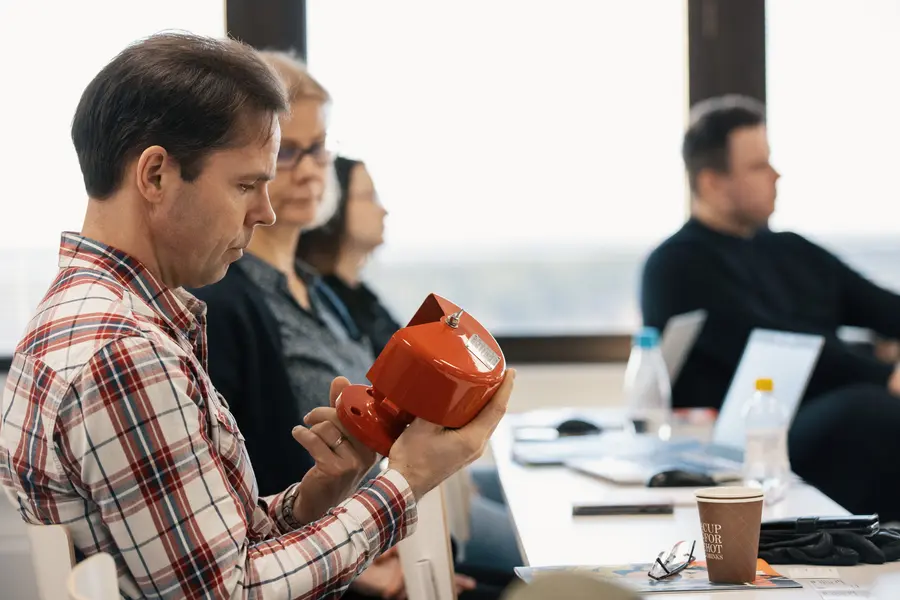 A man seated in a classroom-style setting examines a red industrial device while other attendees sit nearby with notebooks and laptops.
