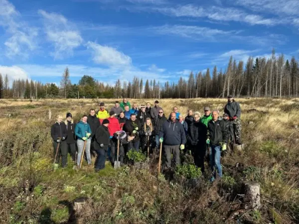 Un numeroso grupo de personas con guantes posa con palas en un campo abierto, con algunos árboles y un cielo azul al fondo.