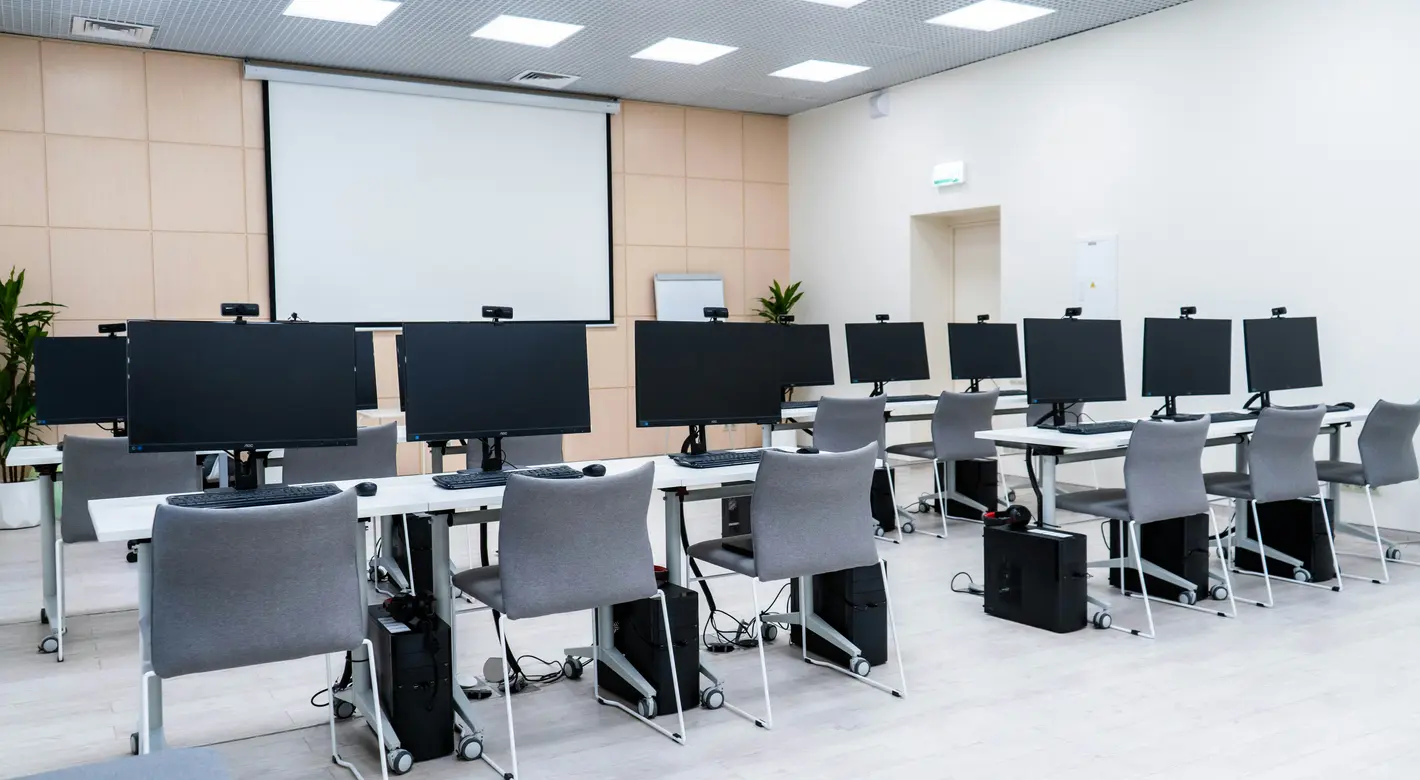 Ein leerer Computerraum mit Reihen von Schreibtischen, Desktop-Monitoren und einer großen Projektionswand an der Vorderseite.