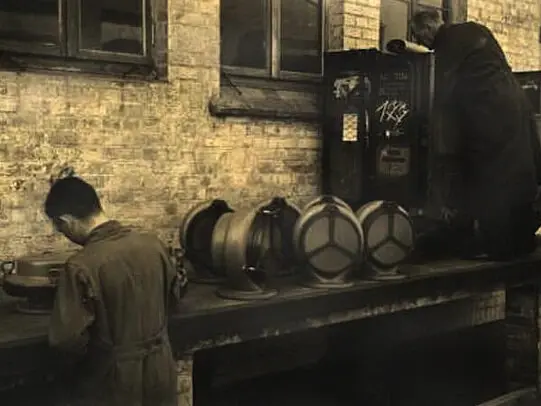 Vintage workshop scene showing workers beside cylindrical metal components arranged on a bench against a brick wall in an industrial setting.