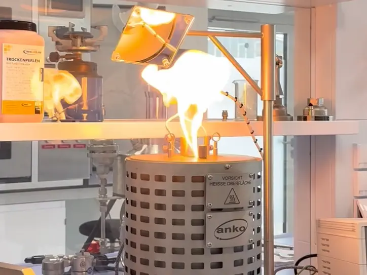 Ein Laborbrenner lodert mit einer leuchtend gelben Flamme über einem Metall-Behälter unter einer montierten Wärmelampe.
