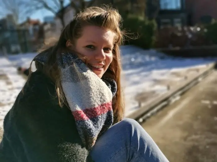 Una mujer sonriente, con una bufanda gruesa a rayas, está sentada al aire libre en un soleado día de invierno, con la nieve y una acera difuminadas al fondo.