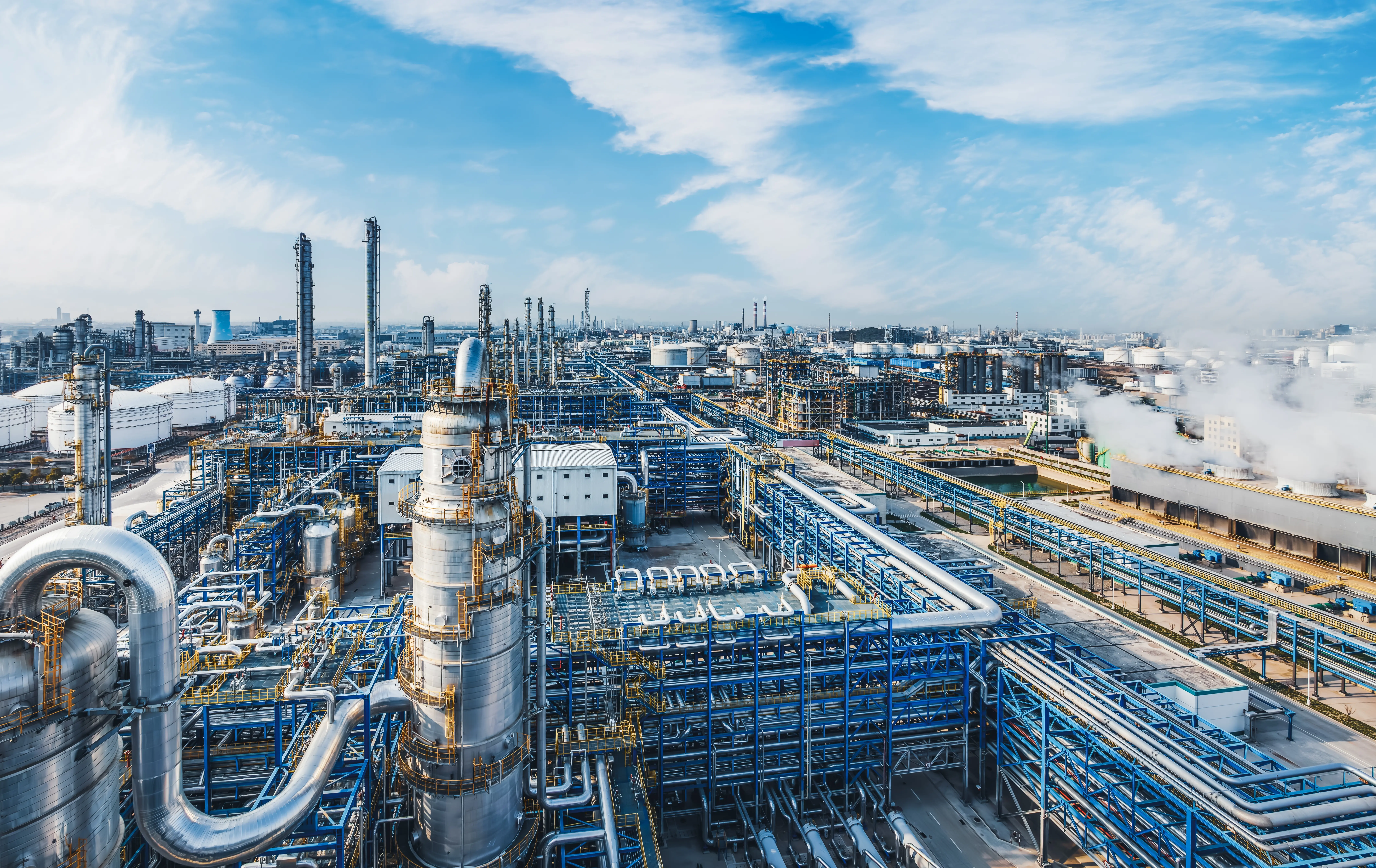 Industrieanlagen, Abgasfackeln und Lagertanks vor blauem Himmel, deren metallische Oberflächen sich darin spiegeln