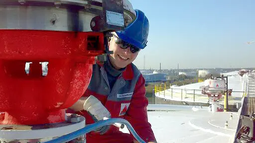 A worker in red coveralls walks through a large industrial plant filled with pipes, valves, and tall processing towers.