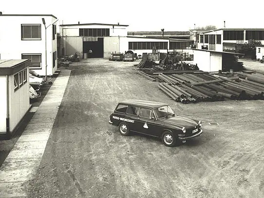 Vintage black-and-white photo of an industrial yard with factory buildings, stacked metal pipes, and a company car parked in the center.