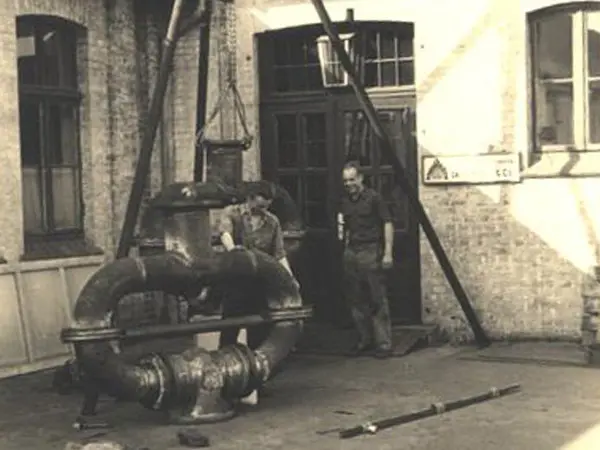 Vintage black-and-white photo of two men standing beside a large industrial pipe assembly being lifted in a brick courtyard.
