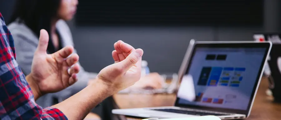 Close-up of a person gesturing with their hands while discussing ideas at a table with an open laptop, with another participant blurred in the background.