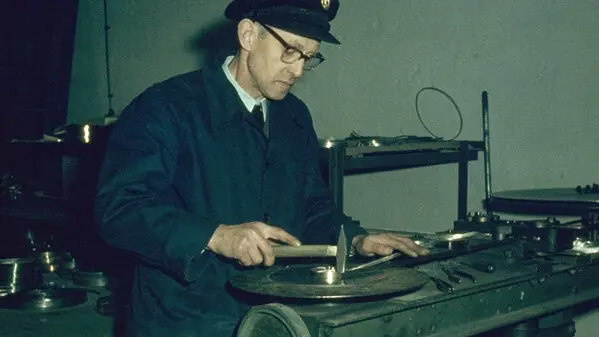 A man in a work coat and cap uses a hand tool at a workbench with metal parts and machinery in a workshop.