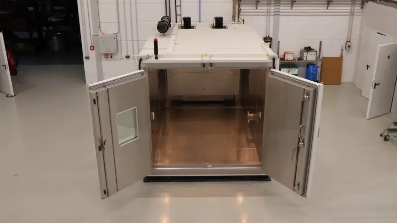 Open industrial test chamber with double doors revealing a stainless steel interior inside a clean factory or laboratory hall.